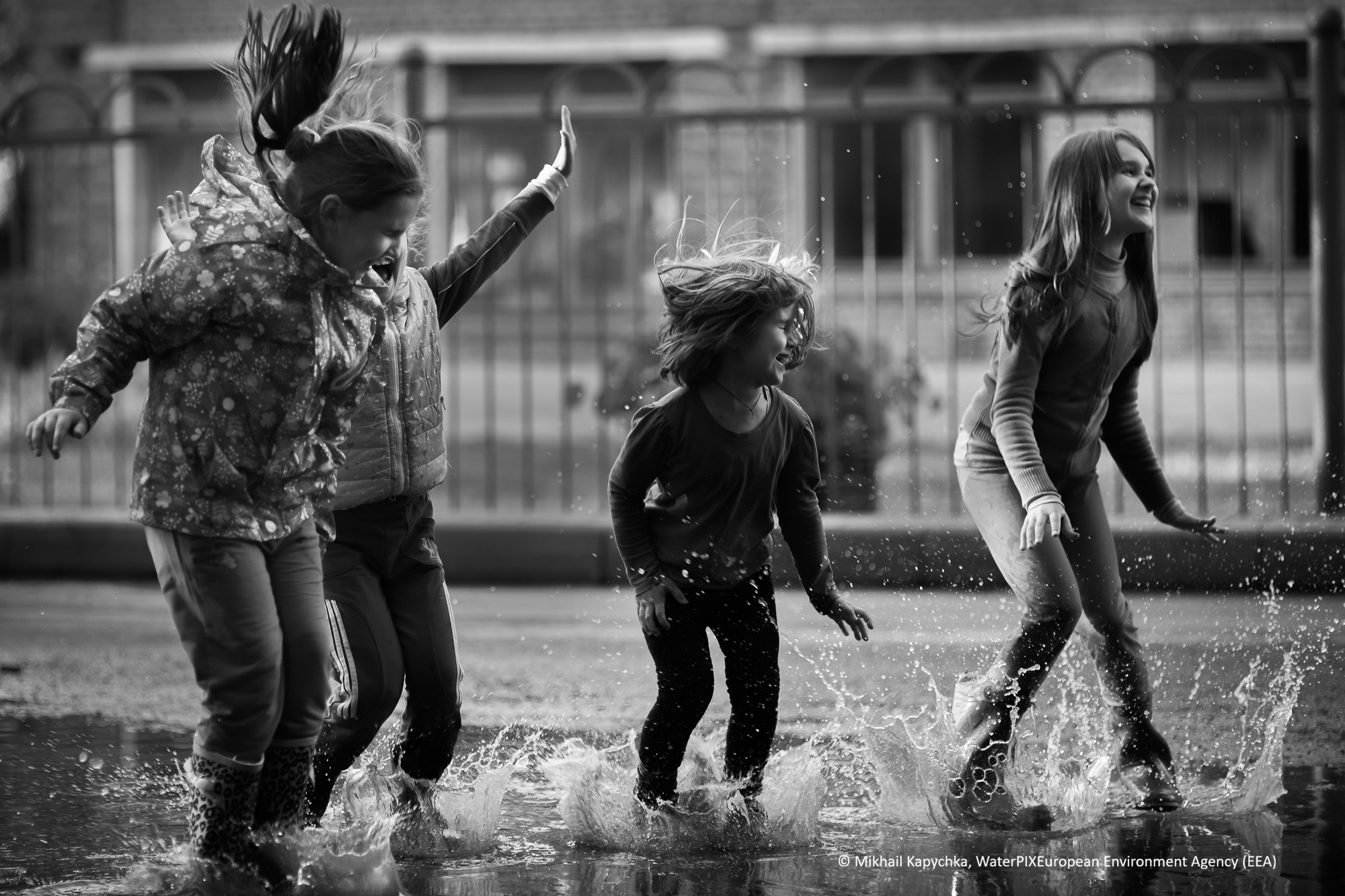 Eine Gruppe von Kindern springt in Pfützen. Das Schwarzweißfoto zeigt einen heiteren Moment des Spielens nach dem Regen.